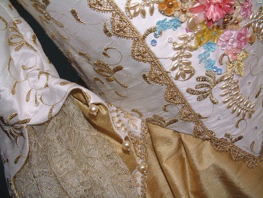 The sleeves, edged in hand-stitched gold pearls, are cut off-the-shoulder for a unique design feature. The boned stomacher is embellished with a variety of flowers: roses, forget-me-nots, and peonies in a variety of colours and fabrics including silk and velvet.  Ivy leaves in a soft green velvet tone beautifully with the gold apron underskirt and gold lace edgings. Tendrils of pearls and scattered crystals ensure a stunning impact.